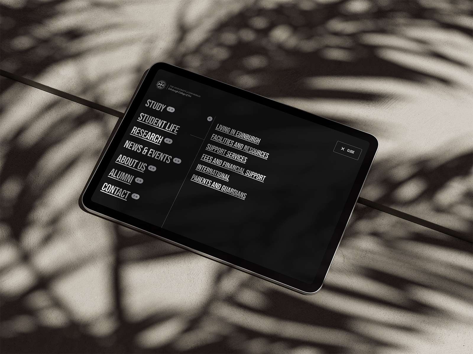 Tablet on a stone surface with shadows, showing the Edinburgh College of Art website menu with study, research, and contact links.